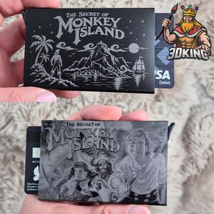 May include: A black metal card holder featuring laser-engraved artwork from The Secret of Monkey Island. One side displays a tropical island scene with a ship, while the other side depicts characters with swords. A small black lever is on the side.