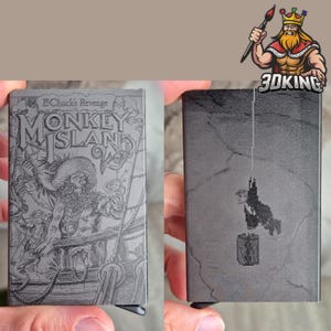 May include: A dark gray metal card holder featuring an engraved Monkey Island 2 LeChuck's Revenge design on one side, showing a pirate captain on a ship. The reverse side displays a pixelated character hanging from a rope above a barrel.
