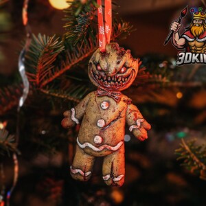 Pode incluir: Um enfeite de Natal de homem de gengibre com um rosto assustador e rachado e uma gravata borboleta vermelha. O enfeite está pendurado em uma árvore de Natal.