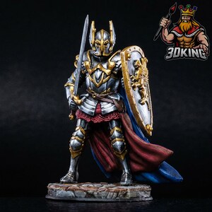 May include: A miniature knight figurine in silver and gold armor, holding a sword and shield. The shield features a golden lion emblem. The knight stands on a stone base, with a red and blue cape. The 3DKING logo is in the upper right corner.