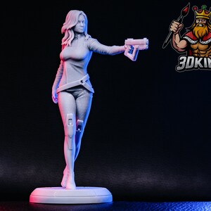 Può includere: Una statuetta bianca stampata in 3D di una donna che tiene una pistola. Indossa una camicia a maniche lunghe e pantaloni. La statuetta è in piedi su una base rotonda.