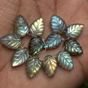 May include: Close-up of a collection of leaf-shaped beads. The beads are carved from a stone with iridescent qualities, displaying shades of green, blue, and yellow. Each bead is strung on a white thread, showcasing the intricate details.