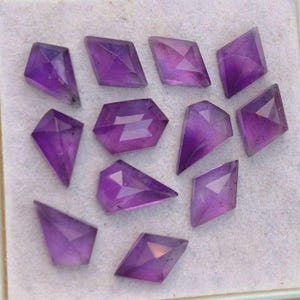 May include: A collection of twelve faceted amethyst gemstones in various shapes and sizes. The gemstones are a deep purple color and have a shiny, polished finish.