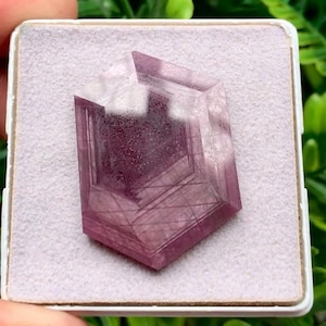 Puede incluir: Una piedra preciosa facetada de forma hexagonal con un tono translúcido púrpura y magenta intenso. La piedra presenta inclusiones lineales internas y una superficie pulida, descansando sobre una base de exhibición cuadrada de color gris claro.