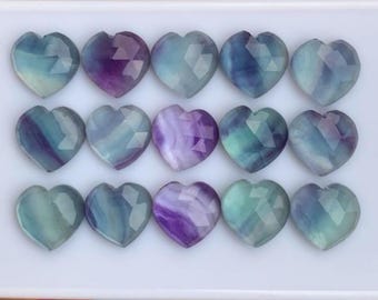 May include: Fifteen heart-shaped gemstones in shades of blue, purple, and green. Each stone has a faceted surface, creating a sparkling effect. The gemstones are arranged in rows on a white surface, showcasing their varying colours and patterns.