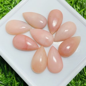 Könnte beinhalten: Eine Sammlung von neun tropfenförmigen rosa Opal-Cabochons. Die Opale sind glatt und poliert und haben einen perlmuttartigen Glanz.