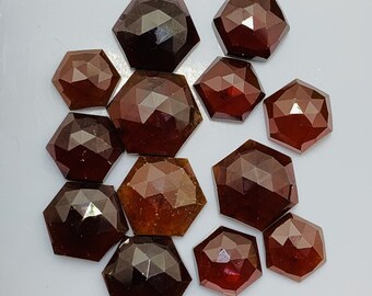 Pode incluir: Uma coleção de 12 gemas facetadas de cor vermelho escuro com forma hexagonal. As gemas estão dispostas sobre uma superfície branca.