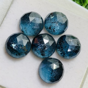 May include: Six round, faceted, blue gemstones with black inclusions. The gemstones are arranged in a cluster on a white surface.