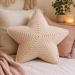 May include: A cream-coloured, star-shaped crochet cushion. The cushion has a textured surface and a circular design in the centre. It is placed on a bed with other cushions and a pink blanket.