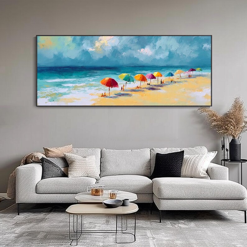 May include: A framed painting of a beach scene. The artwork shows a sunny beach with colourful umbrellas, people, and the ocean. The sky is blue and white, and the sand is yellow. The painting is displayed above a grey sofa.
