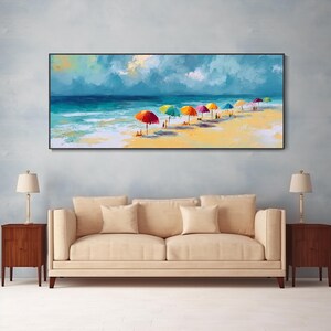 May include: A framed painting of a beach scene. The artwork shows a sunny beach with colourful umbrellas, people, and the ocean under a cloudy sky. The painting is displayed above a beige sofa with two end tables and lamps.