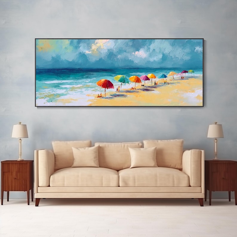 May include: A framed painting of a beach scene. The artwork shows a sunny beach with colourful umbrellas, people, and the ocean under a cloudy sky. The painting is displayed above a beige sofa with two end tables and lamps.