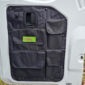 May include: Black fabric organiser with multiple pockets attached to the inside of a white car door. The organiser has a green logo with the text "G.I. Glamping" and "Leighoom, Co. UK".