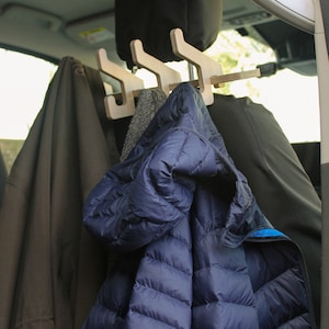 May include: Wooden car headrest coat rack with three hooks, holding a navy blue puffer jacket, a grey scarf, and a brown coat. The rack is designed to hang clothing in a vehicle.