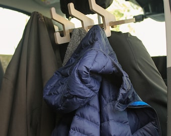 May include: Wooden car headrest coat rack with three hooks, holding a navy blue puffer jacket, a gray scarf, and a brown coat. The rack is designed to hang clothing in a vehicle.