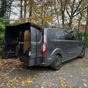 May include: A grey van with the rear doors open and a black canvas awning attached to the roof. The van is parked on a gravel driveway with fallen leaves.