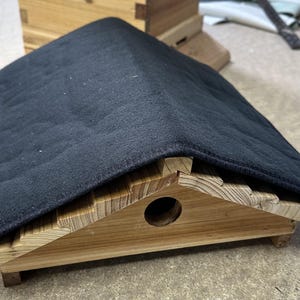 May include: A wooden birdhouse with a dark gray, quilted roof. The birdhouse has a triangular shape with a round entry hole. The wooden structure is made of light-colored wood.