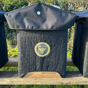 Pode incluir: Três colmeias pretas com uma base de madeira e uma cobertura de tecido preto. As colmeias têm um desenho de nó celta dourado com o texto "Celtic Bees" na frente.