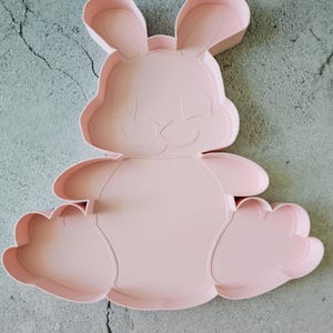 Puede incluir: Cortador de galletas de plástico rosa en forma de conejo sentado. El cortador tiene un diseño detallado con una cara, orejas y patas.