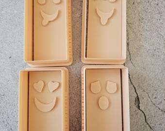 Könnte beinhalten: Vier hellpfirsichfarbene rechteckige Ausstecher mit verschiedenen Emoji-Gesichtern. Die Emojis umfassen ein lächelndes Gesicht mit herausgestreckter Zunge, ein zwinkerndes Gesicht mit herausgestreckter Zunge, ein lächelndes Gesicht mit Herzen als Augen und ein neutrales Gesicht.