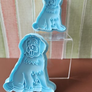 Puede incluir: Dos cortadores de galletas de perro de plástico azul claro. Los cortadores tienen la forma de un perro sentado con una cola esponjosa.