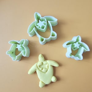 May include: Four sea turtle-shaped cookie cutters and a yellow cookie on a yellow background. The cookie cutters are light green plastic with a star design on the turtle's shell. The cookie is yellow and has the same star design.