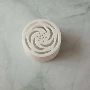 Op de afbeelding: Ronde, witte luchtverfrisser met een swirl-ontwerp en kleine gaatjes voor geurvrijgave.