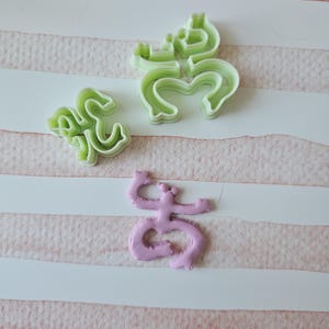 May include: Three cookie cutters in the shape of a stylized letter "S". Two are green plastic and one is pink plastic.