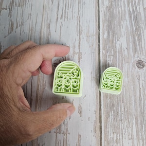 May include: Two light green cookie cutters. The larger one is held by a hand. Both cutters are rectangular with rounded tops and feature a design of three figures with star and leaf details. The cutters are on a light wood surface.