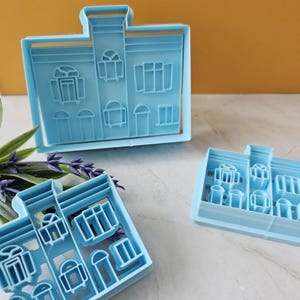 Può includere: Tre tagliabiscotti in plastica blu a forma di case con diversi disegni di finestre e porte.