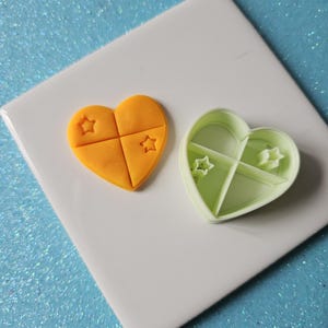 May include: An orange heart-shaped cookie with star imprints, next to a green heart-shaped cookie cutter. The cookie is divided into four sections. The cutter is divided into four sections, each with a star-shaped imprint.
