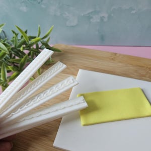Puede incluir: Cuatro herramientas de decoración de pasteles de fondant de silicona blanca con diferentes patrones en relieve. Las herramientas están sobre una superficie blanca con un cuadrado de fondant amarillo.