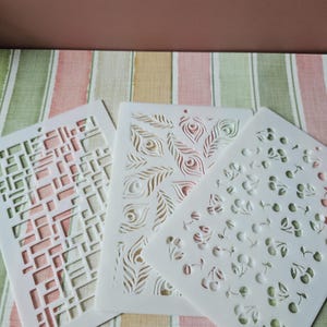 May include: Three white plastic stencils with different designs. The first stencil has a geometric pattern of squares and rectangles. The second stencil has a peacock feather design. The third stencil has a repeating pattern of cherries and leaves.