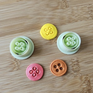 Peut inclure: Cinq emporte-pièces en forme de bouton de différentes couleurs : jaune, rose et orange, et deux découpoirs vert clair. Les découpoirs sont disposés sur une surface en bois brun clair. Les boutons ont quatre trous.