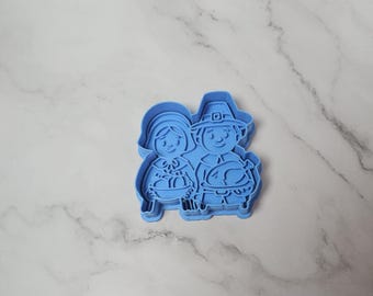 May include: A blue plastic cookie cutter featuring a cartoon couple, a man and a woman, dressed in traditional Thanksgiving attire. The man is holding a turkey. The cutter is designed for making festive cookies.