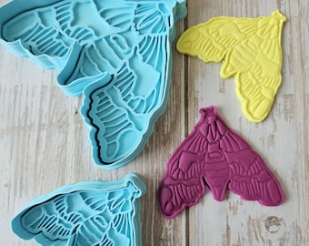 May include: A collection of moth-shaped cookie cutters in various colors. Two light blue cutters are next to a yellow and a magenta moth-shaped cookie. The cutters have detailed wing patterns, perfect for baking themed treats.