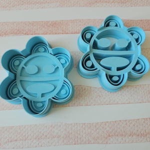 May include: Two light blue plastic cookie cutters shaped like a stylized sun with a face. The cutters are on a white and pink striped surface.