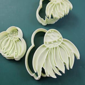 May include: Three light green plastic flower-shaped cookie cutters. Each cutter has a detailed flower design with petals and a central disc. The cutters are arranged on a dark green surface.