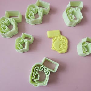 May include: A set of seven green plastic cookie cutters shaped like a cartoon animal. The cutters are stacked on top of each other. There is also a yellow piece of fondant shaped like the cartoon animal.