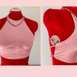 May include: A pink halter top with a silver chain necklace featuring a heart pendant. The top is made of a silky fabric and has a tie closure in the back.