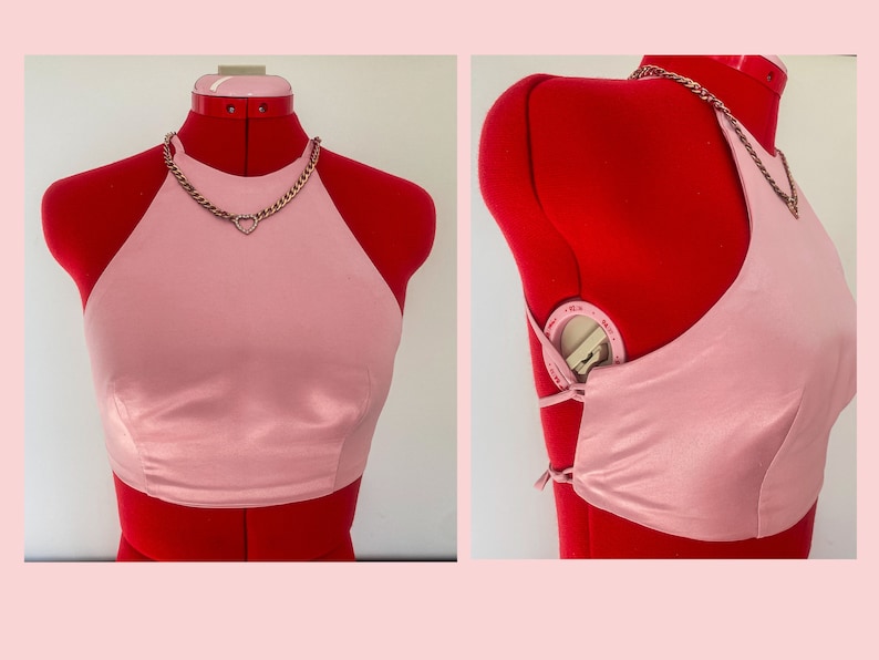 May include: A pink halter top with a silver chain necklace featuring a heart pendant. The top is made of a silky fabric and has a tie closure in the back.