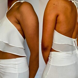 May include: A two-piece white outfit featuring a halter top with layered ruffles and a matching skirt. The top ties at the neck and back. The outfit is made of a lightweight fabric, perfect for warm weather.