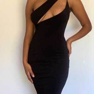 May include: A black, one-shoulder dress with a cutout detail. The dress has a fitted silhouette and a thin strap on one shoulder. The fabric appears to be a smooth, stretchy material.