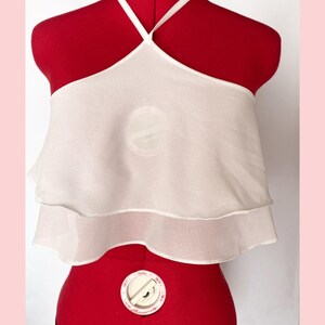May include: A white halter top with a layered design. The top is made of a sheer, lightweight fabric and features a halter neckline with thin straps. The garment is displayed on a red dress form.