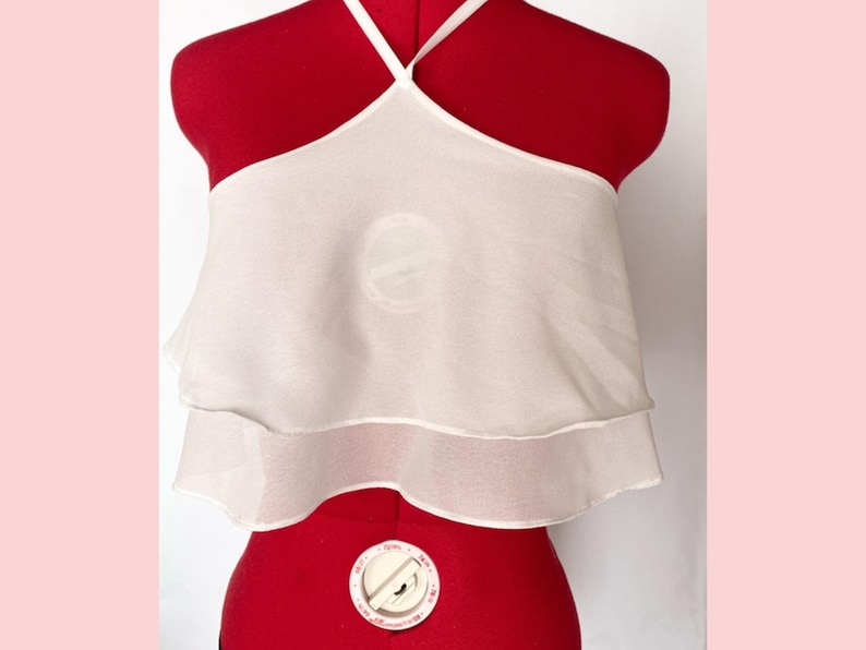 May include: A white halter top with a layered design. The top is made of a sheer, lightweight fabric and features a halter neckline with thin straps. The garment is displayed on a red dress form.