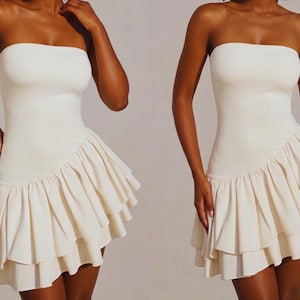 May include: Off-white strapless dress with a fitted bodice and a layered, ruffled skirt. The dress has a simple, elegant design, perfect for a special occasion. The fabric appears to be a soft, flowing material.