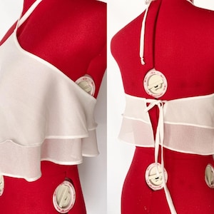 May include: A white halter top with layered ruffles. The top is displayed on a red dress form. The back features a tie closure and a halter neck strap. The fabric appears to be a lightweight, sheer material.