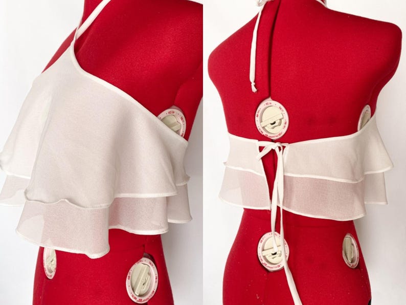 May include: A white halter top with layered ruffles. The top is displayed on a red dress form. The back features a tie closure and a halter neck strap. The fabric appears to be a lightweight, sheer material.
