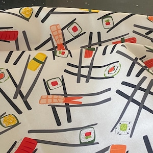 May include: White fabric with a repeating pattern of sushi rolls, nigiri, and shrimp. The sushi is in various colors, including red, orange, yellow, and green. Black lines and chopsticks are also part of the design.