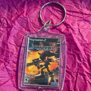 May include: A clear acrylic keyring featuring the cover art from the PlayStation 2 game "Shadow the Hedgehog." The artwork shows the character Shadow with a weapon against a fiery background. The keyring has a silver metal ring.